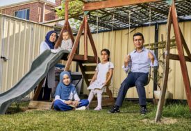 A family with three children sit outside in a backyard on a swing set