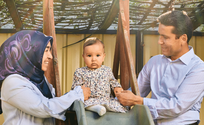 A woman wearing a headscarf and a man hold their baby who is sitting on a slide in their backyard.
