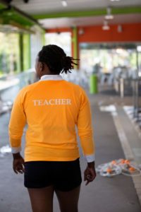 A woman standing - the back of her shirt says 'teacher'.