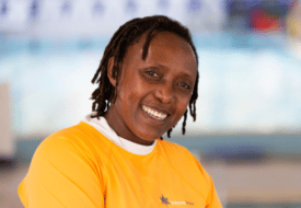 A woman with short dreadlocks smiles at the camera. She is wearing a yellow top.