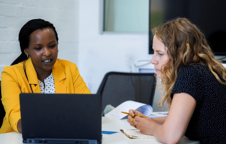 Two women sit in discussion in front of a laptop computer. There is also paperwork and a notebook on the table