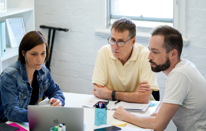 Three people sit around a table at the Asylum Seekers Centre, looking seriously at a laptop