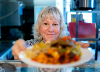 A woman smiles as she passes a plate of colourful food toward the camera