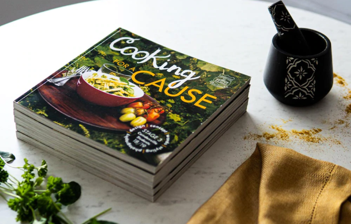 A stack of cookbooks titled 'Cooking for a Cause' sit on a table next to a napkin, herbs and mortar and pestle