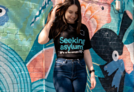 A woman wearing an Asylum Seekers Centre tshirt stands in front of a brightly patterned mural