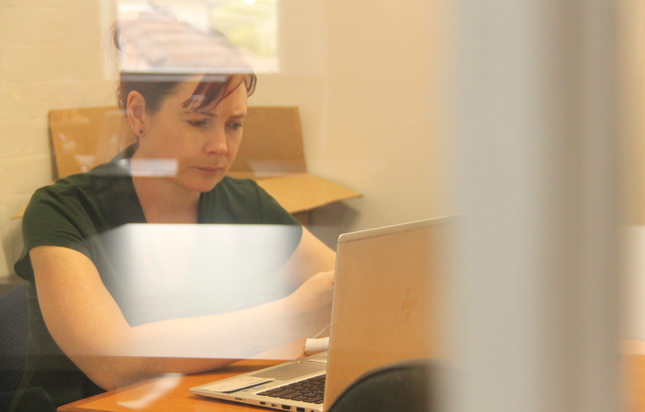 Andrea is shown sitting at a table looking at a laptop. The photo is taken through a glass door.