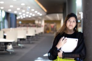 A young woman holds a laptop in a university library.