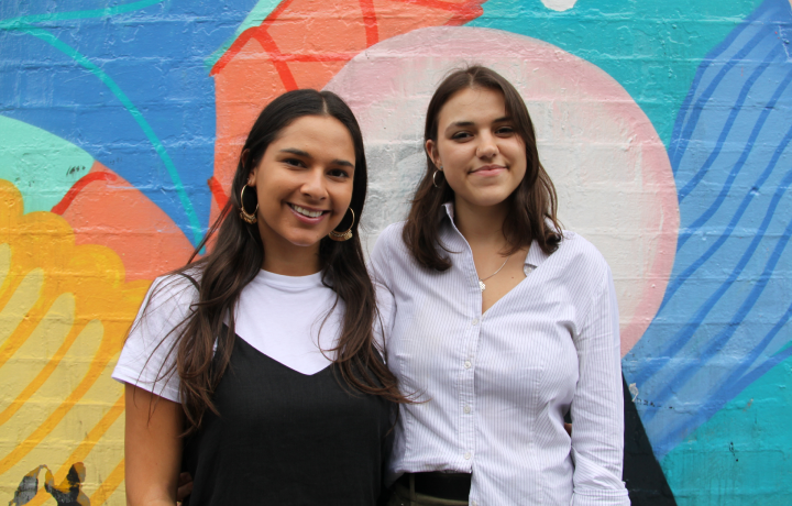 Two students, Lyla and Sureesha, stand in front of a colourful mural smiling at the camera