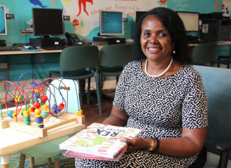 A woman sits in front of computers at the Asylum Seekers Centre. She is wearing a black and white dress and holds picture books