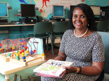 A woman sitting down next to children's toys, holding a children's book and smiling