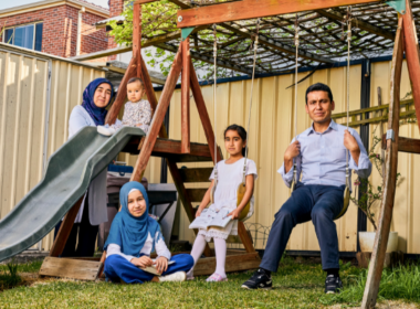 A family - a woman, baby, two girls and a man -poses in their backyard on a swing set and slide. The woman and eldest daughter wear Muslim headscarves.