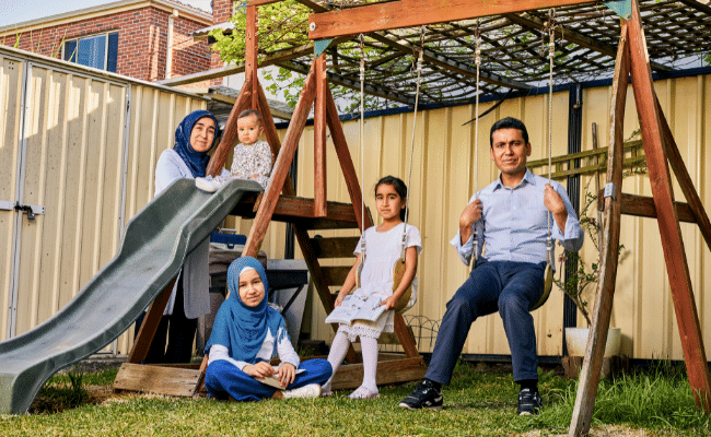 A family - a woman, baby, two girls and a man -poses in their backyard on a swing set and slide. The woman and eldest daughter wear Muslim headscarves.