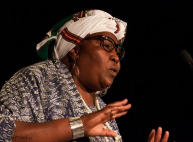 A woman speaking at an event, wearing an African headdress.