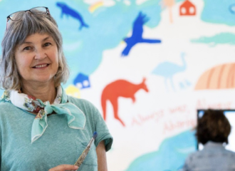 A woman stands in front of a brightly coloured mural. She is wearing a blue top and has a scarf tied around her neck.