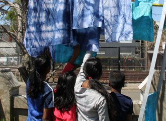 Children stand with their backs to the camera. They are outside and looking at some shibori dyed fabric that is hanging above.