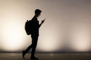 A silhouette of a man walking with a backpack and looking at his mobile phone