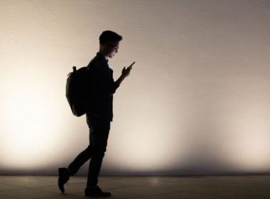 A silhouette of a man walking with a backpack and looking at his mobile phone