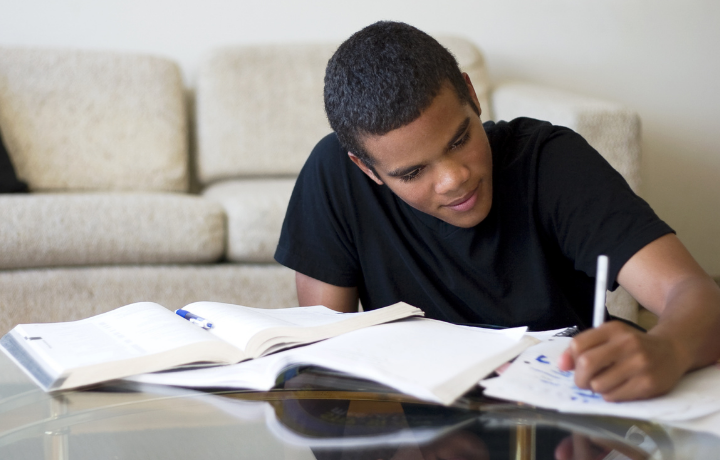 A man in a black shirt sits in front of a white couch. He is writing in a notebook on the coffee table in front of him.