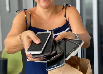 A woman is holding six various mobile phones and passing them toward the camera