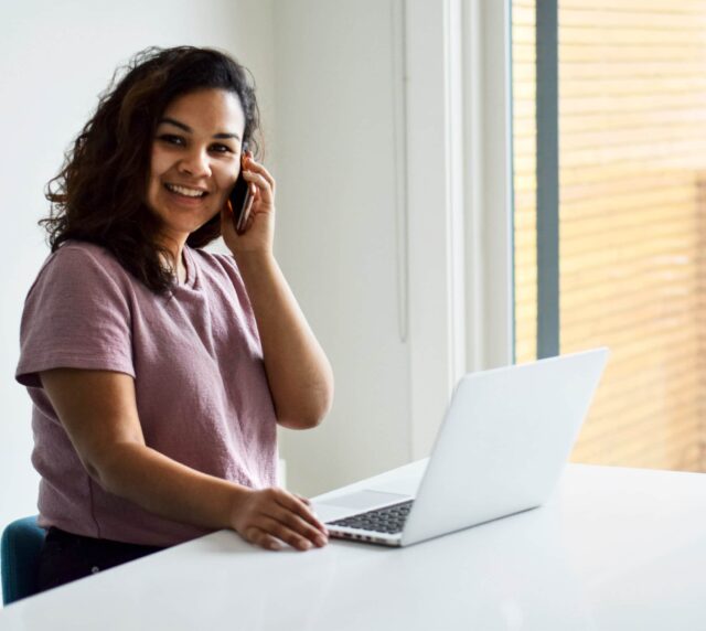 A woman smiles while chatting on the phone and using a laptop.