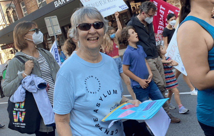 A woman wearing an Asylum Seekers Centre tshirt walks in a rally, smiling and holding flyers.