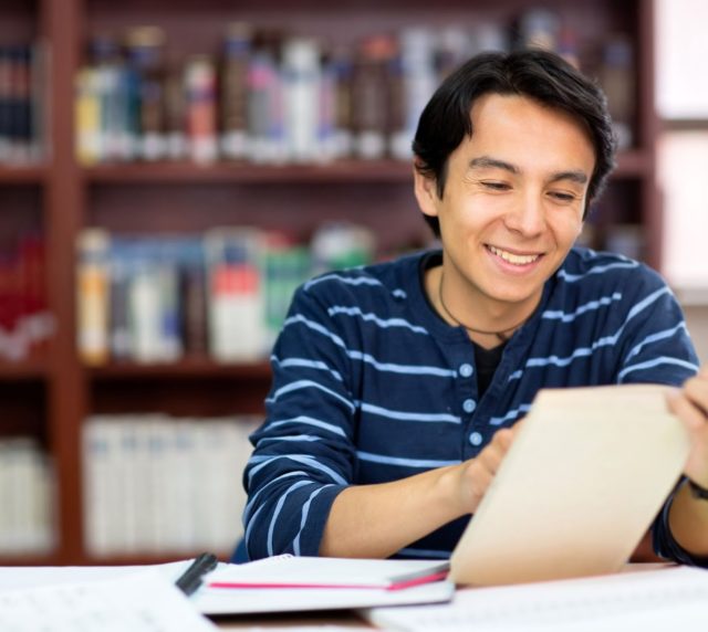 A young man wearing a blue shirt sits in a library with a pile of books in front of him. He is holding a book open and smiling.