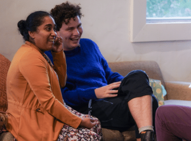 Two people sit on a couch, chatting and smiling, at the Asylum Seekers Centre's National Volunteer Week event.