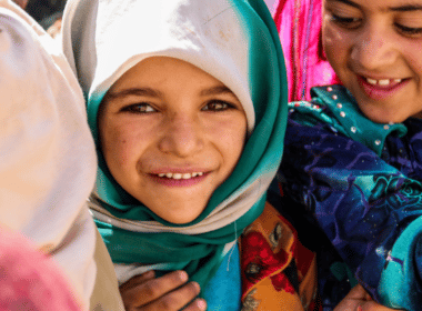 A young girl wearing a headscarf looks at the camera, she is surrounded by other children wearing colourful clothes.