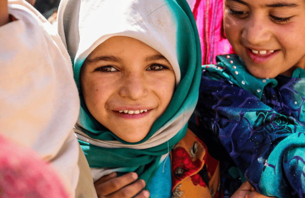 A young girl wearing a headscarf looks at the camera, she is surrounded by other children wearing colourful clothes.