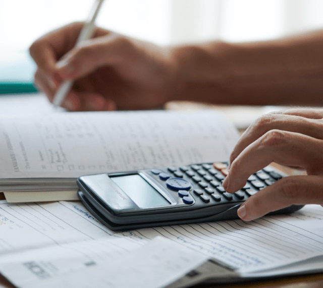 A close-up of a person's hands using a calculator and writing in a notebook with a pen.