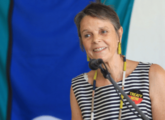 An ASC community speaker talks with a microphone, she is wearing a badge that says 'Treaty now'.