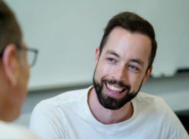 A man with a beard smiles while speaking to another man.
