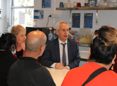 A man, Minister Andrew Giles, sits at a table and speaks to a group of people who are supported by the Asylum Seekers Centre
