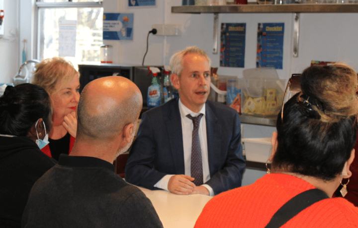 A man, Minister Andrew Giles, sits at a table and speaks to a group of people who are supported by the Asylum Seekers Centre