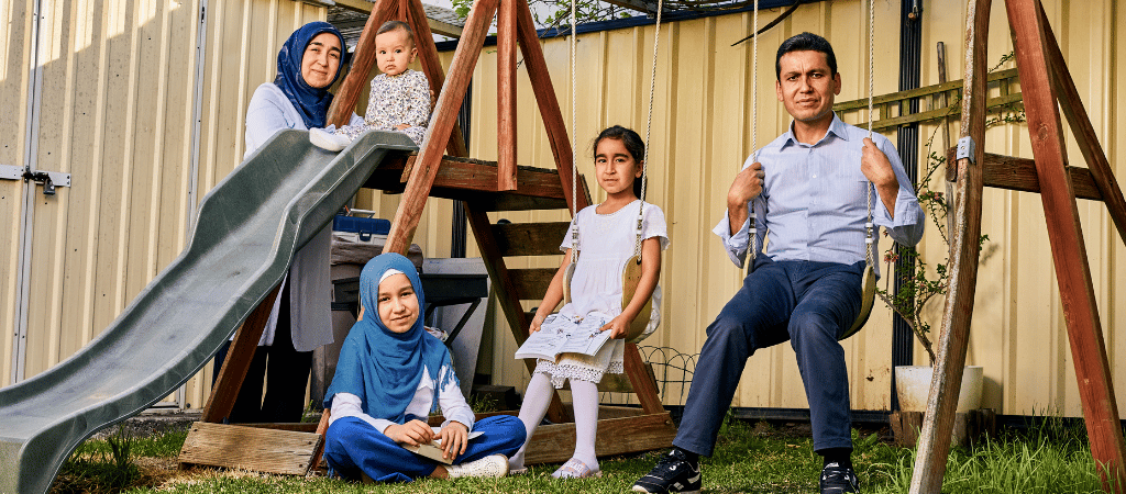 A family - a woman, baby, two girls and a man -poses in their backyard on a swing set and slide. The woman and eldest daughter wear Muslim headscarves.