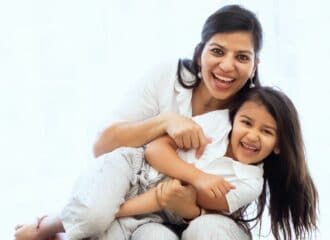 A seated woman holds her young daughter on her lap - they are both smiling and laughing.