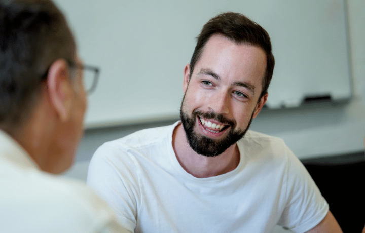 A man with a beard wearing a white shirt smiles as he talks to another person.