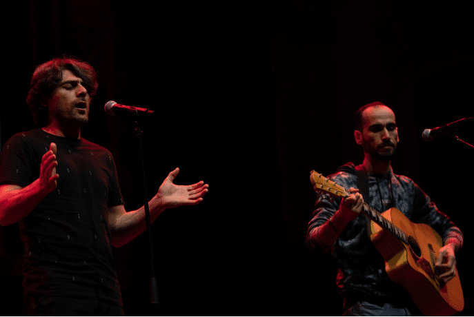Two musicians performing on a stage. One singing and one playing guitar.